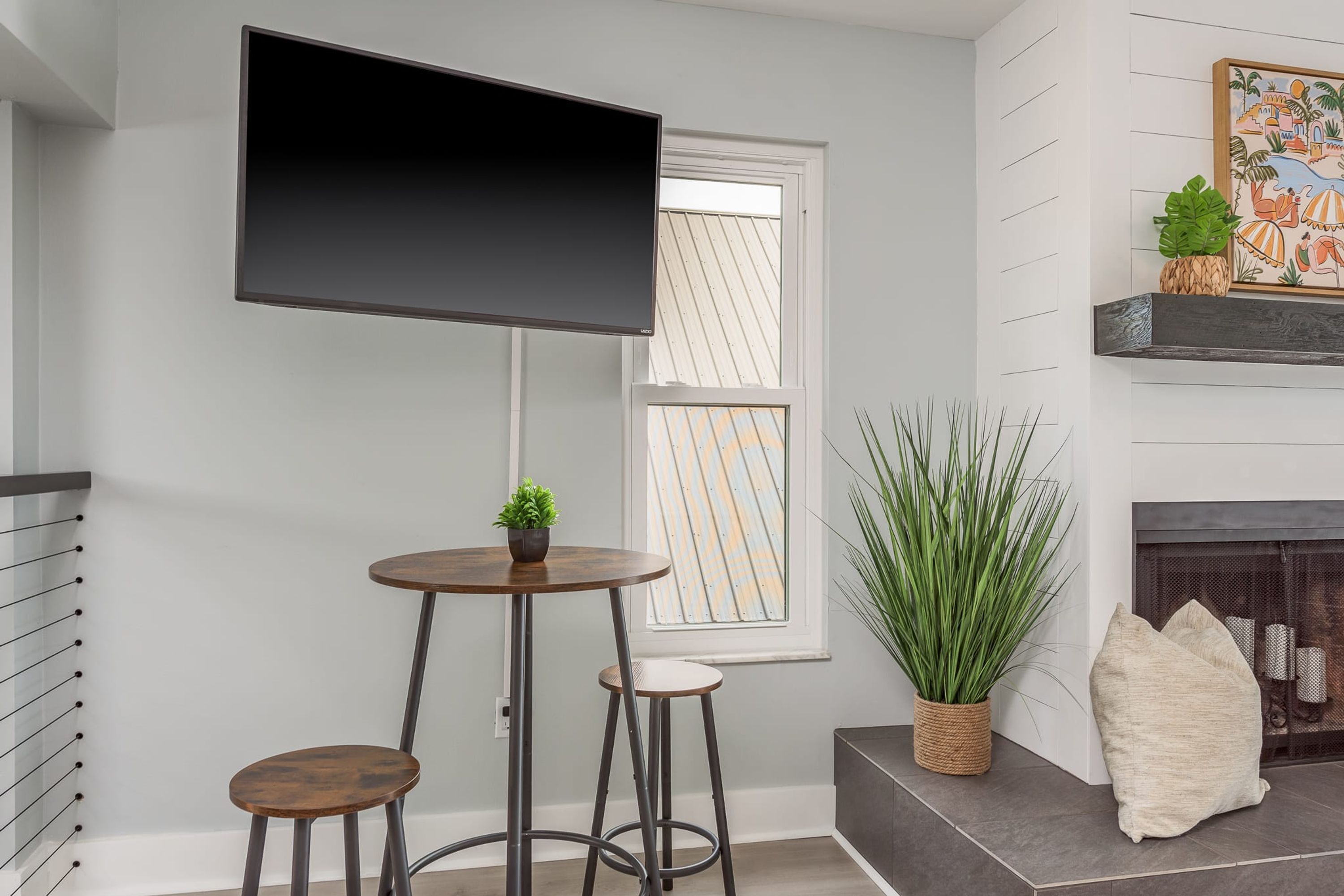 A modern living room with a flat-screen TV mounted on the wall, a round wooden table with two stools, a large potted plant, and a fireplace with decorative pillows.