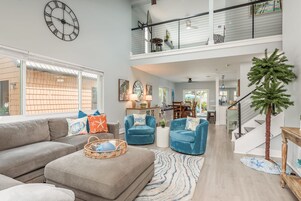 A spacious living room with a grey sofa, blue armchairs, and a coffee table. A large wall clock and a staircase with a railing are visible.