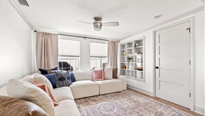 Una sala de estar con un sofá modular, un ventilador de techo, un amplio ventanal con cortinas y una estantería empotrada.