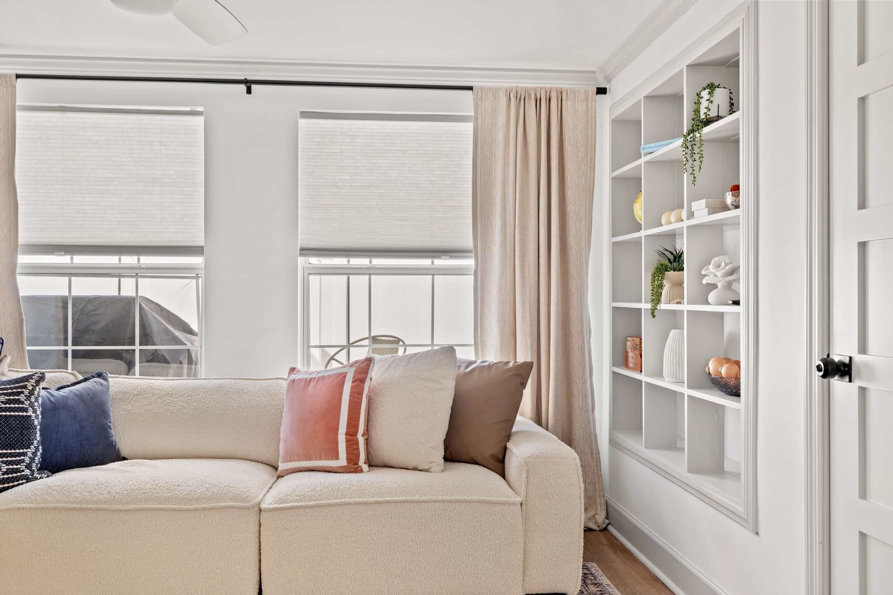A living room with a beige sofa, a white cabinet with shelves, and a window with blinds.