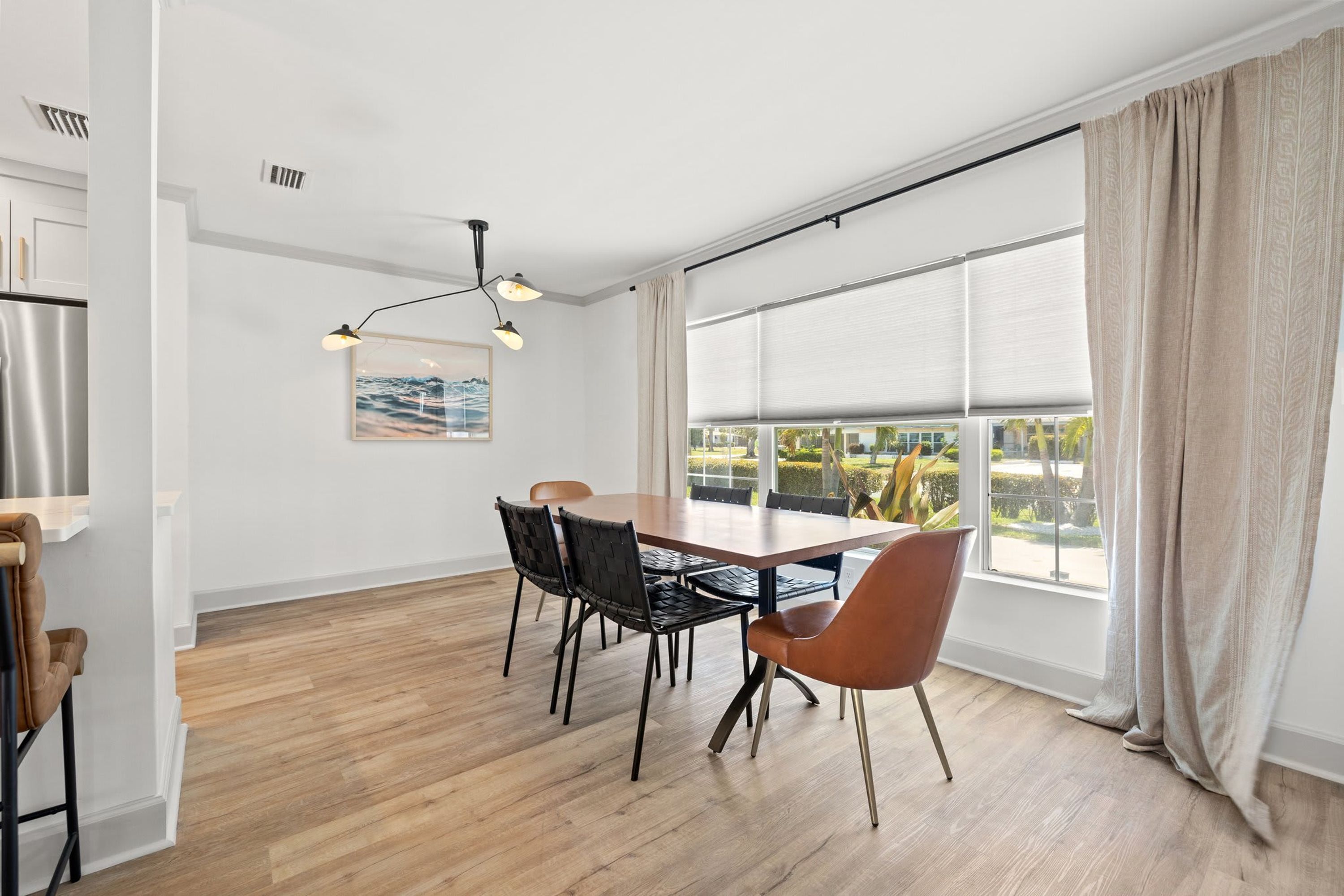 A modern dining area with a wooden table, black and brown chairs, a hanging light fixture, and a large window with blinds.