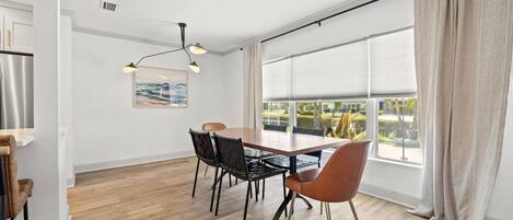 A modern dining area with a wooden table, black and brown chairs, a hanging light fixture, and a large window with blinds.