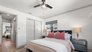 Un dormitorio con una cama grande, un ventilador de techo, una mesita de noche con lámpara y vistas al exterior a través de una ventana.
