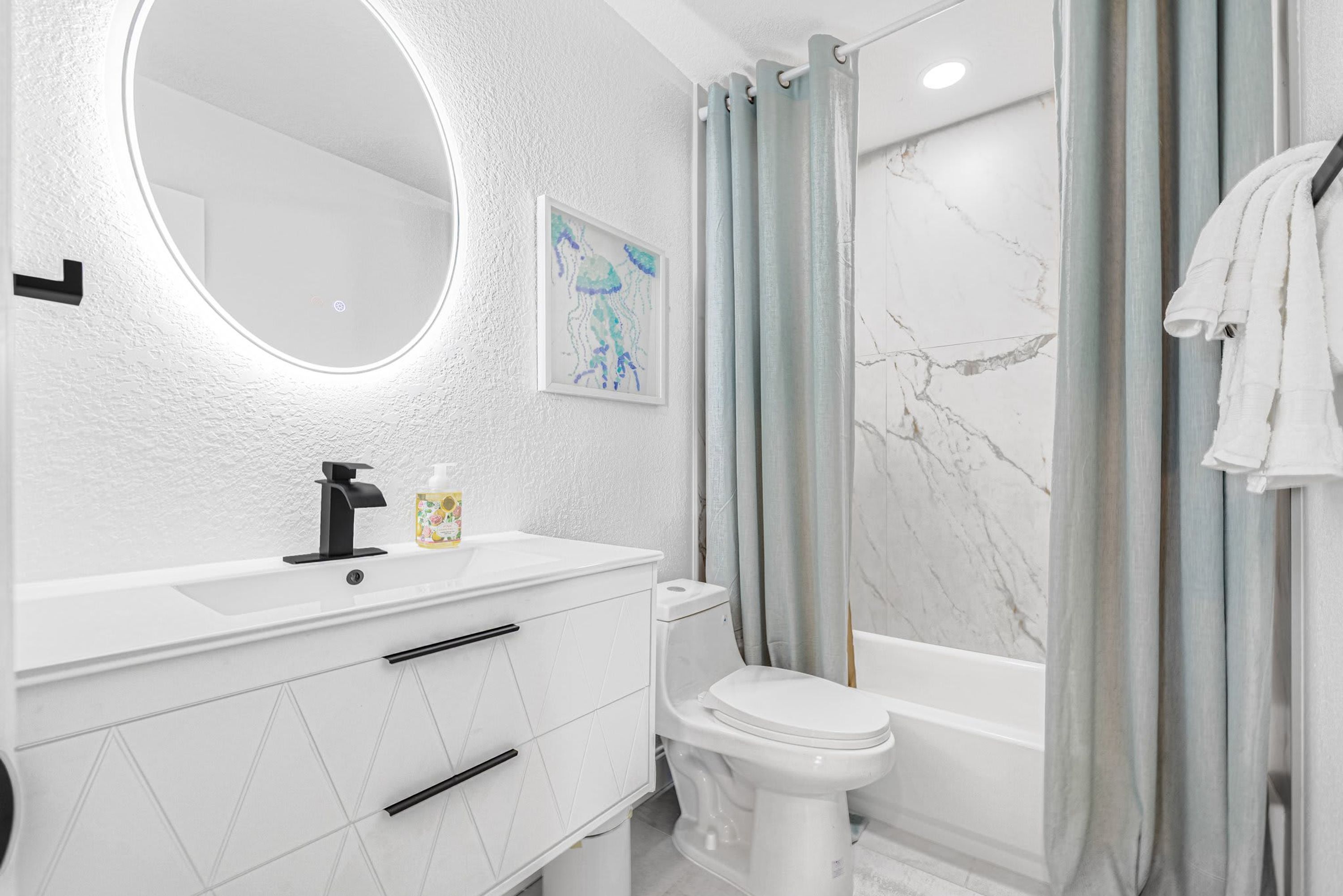 Une salle de bain moderne avec un meuble-lavabo blanc, un miroir rond, des toilettes et une baignoire.