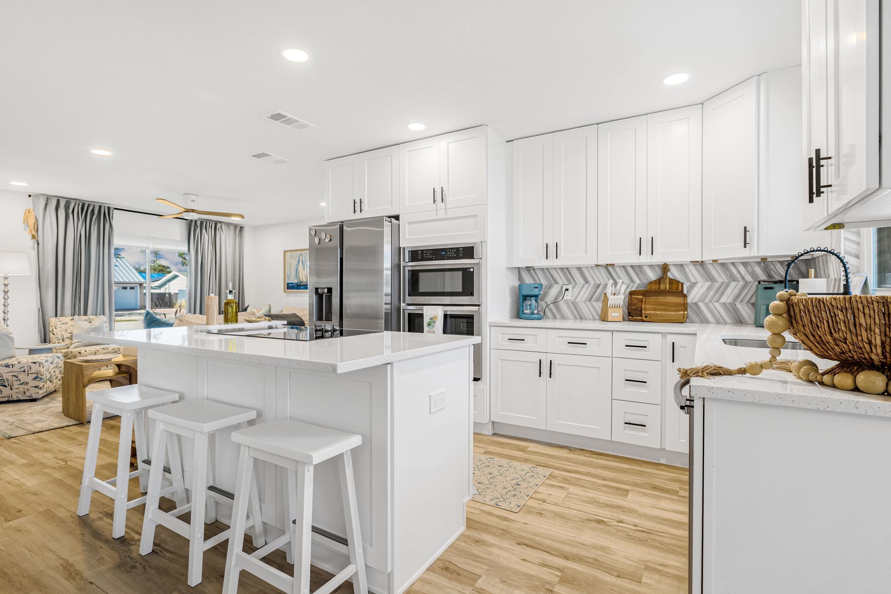 A modern kitchen with white cabinetry, a central island with stools, stainless steel appliances, and a herringbone-patterned backsplash.