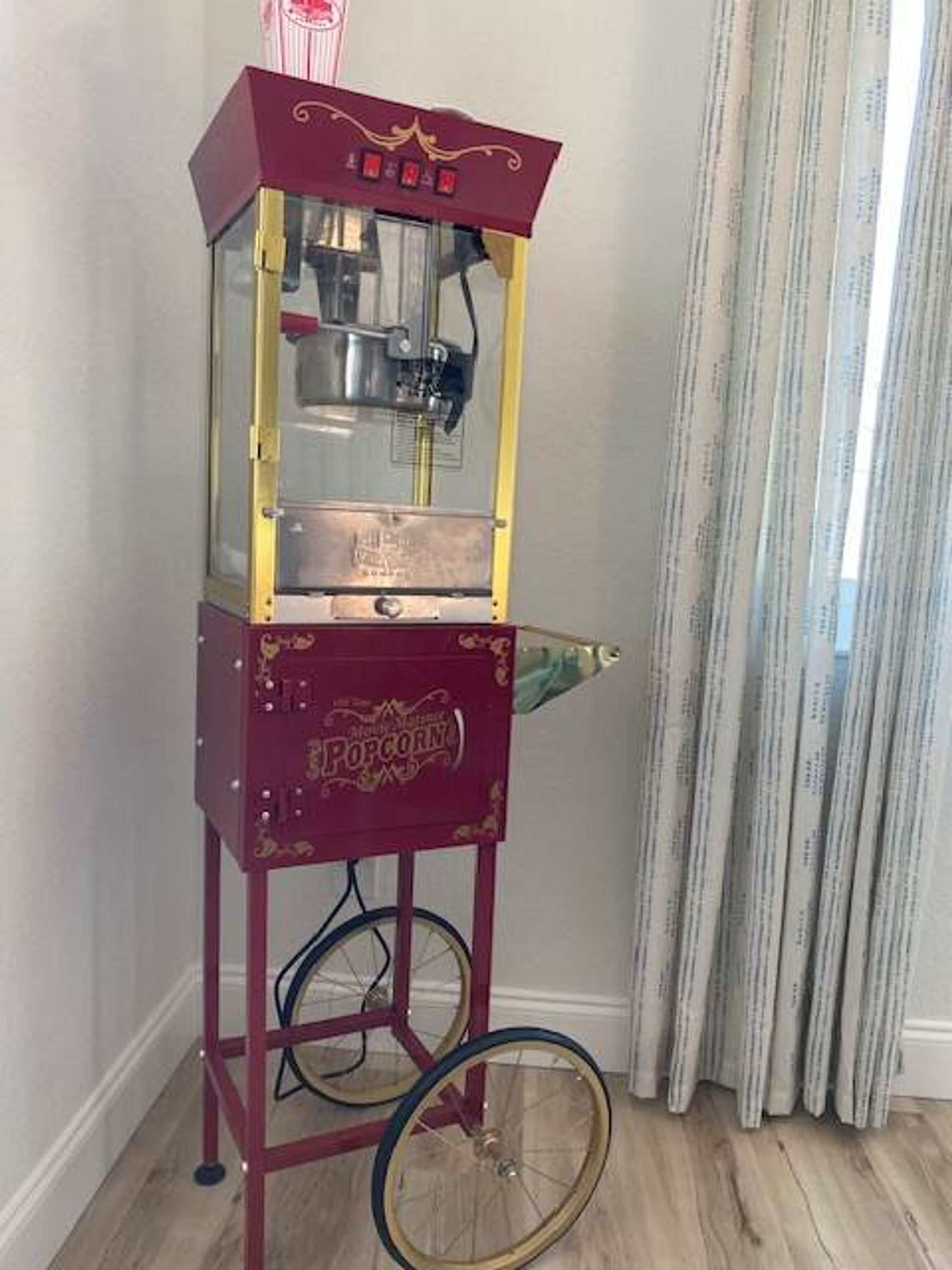 A vintage popcorn machine with a red and gold design, placed against a wall with a curtain.