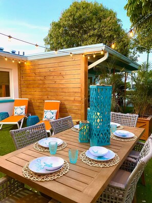 Een terras buiten met een houten eettafel, omringd door rieten stoelen en blauwe kussens.