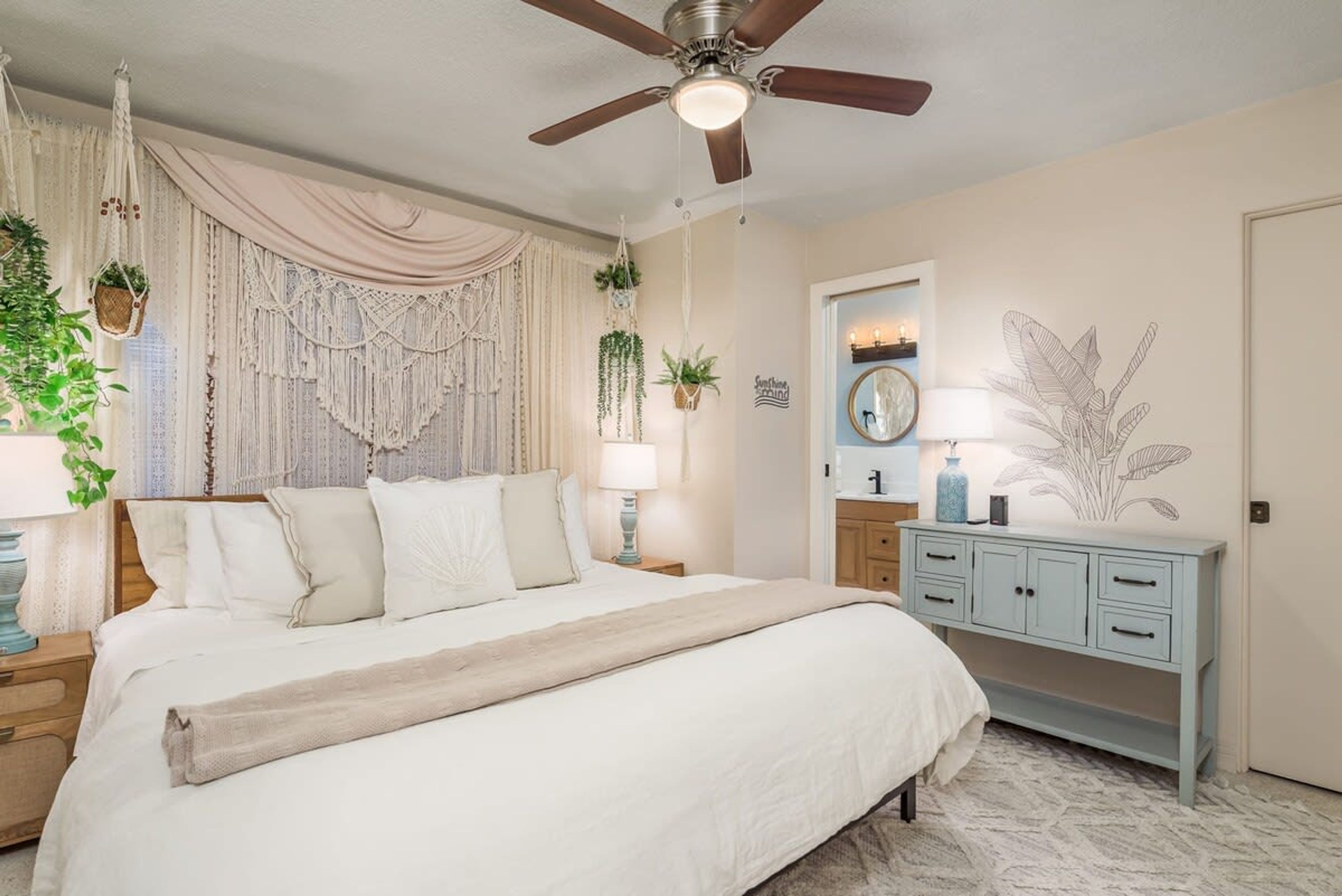 Una camera da letto con un letto, una ventola a soffitto, un comodino con una lampada, una cassettiera e decorazioni a parete tra cui lavori in macramè e piante.