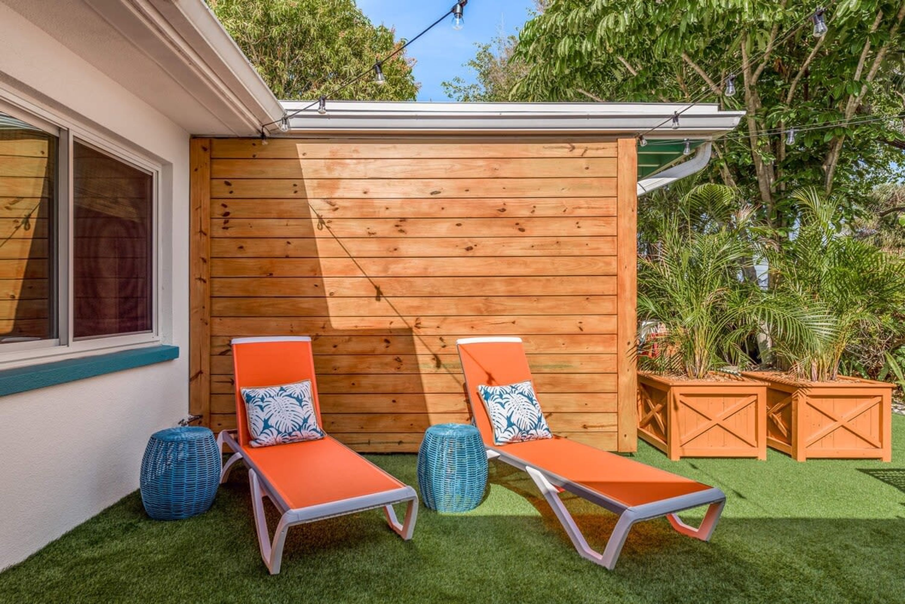Ein kleiner Außenbereich mit zwei orangefarbenen Liegestühlen, einer Holzverkleidung und einem Garten mit Kunstgrashalm und Topfpflanzen.