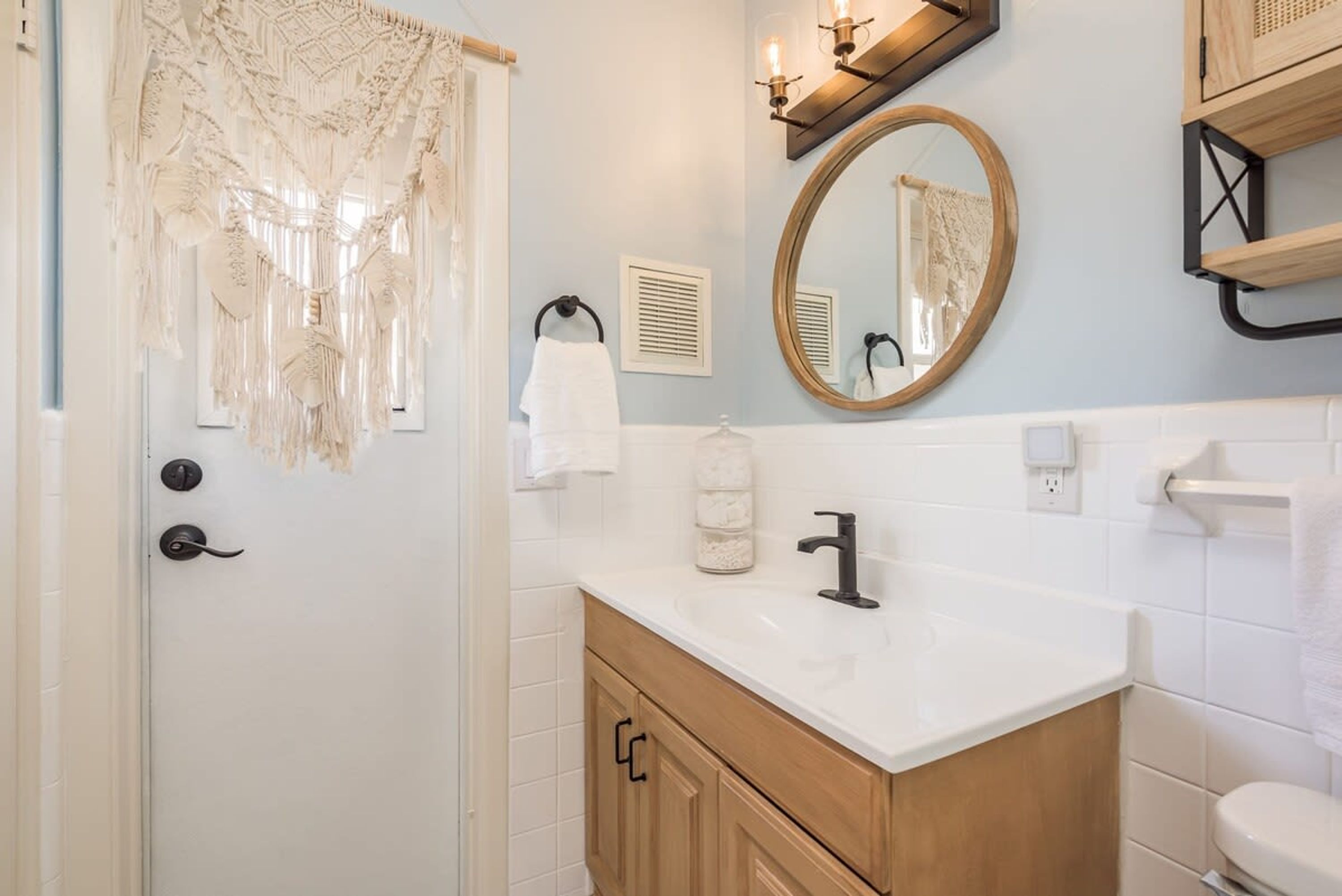 A bathroom with a white sink, wooden vanity, a round mirror, and a decorative macrame curtain.