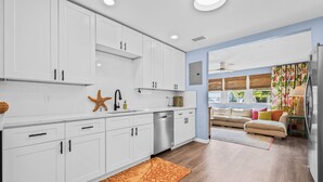 A modern kitchen with white cabinets, a dishwasher, and a starfish decoration. There is a wooden floor, a sofa with colorful cushions, and a window with blinds.