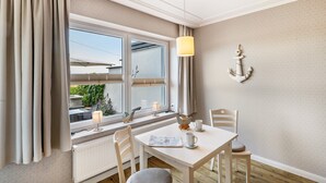 Une pièce comprenant une table, des chaises, une fenêtre avec des rideaux, une lampe et un élément décoratif en forme d’ancre fixé au mur.