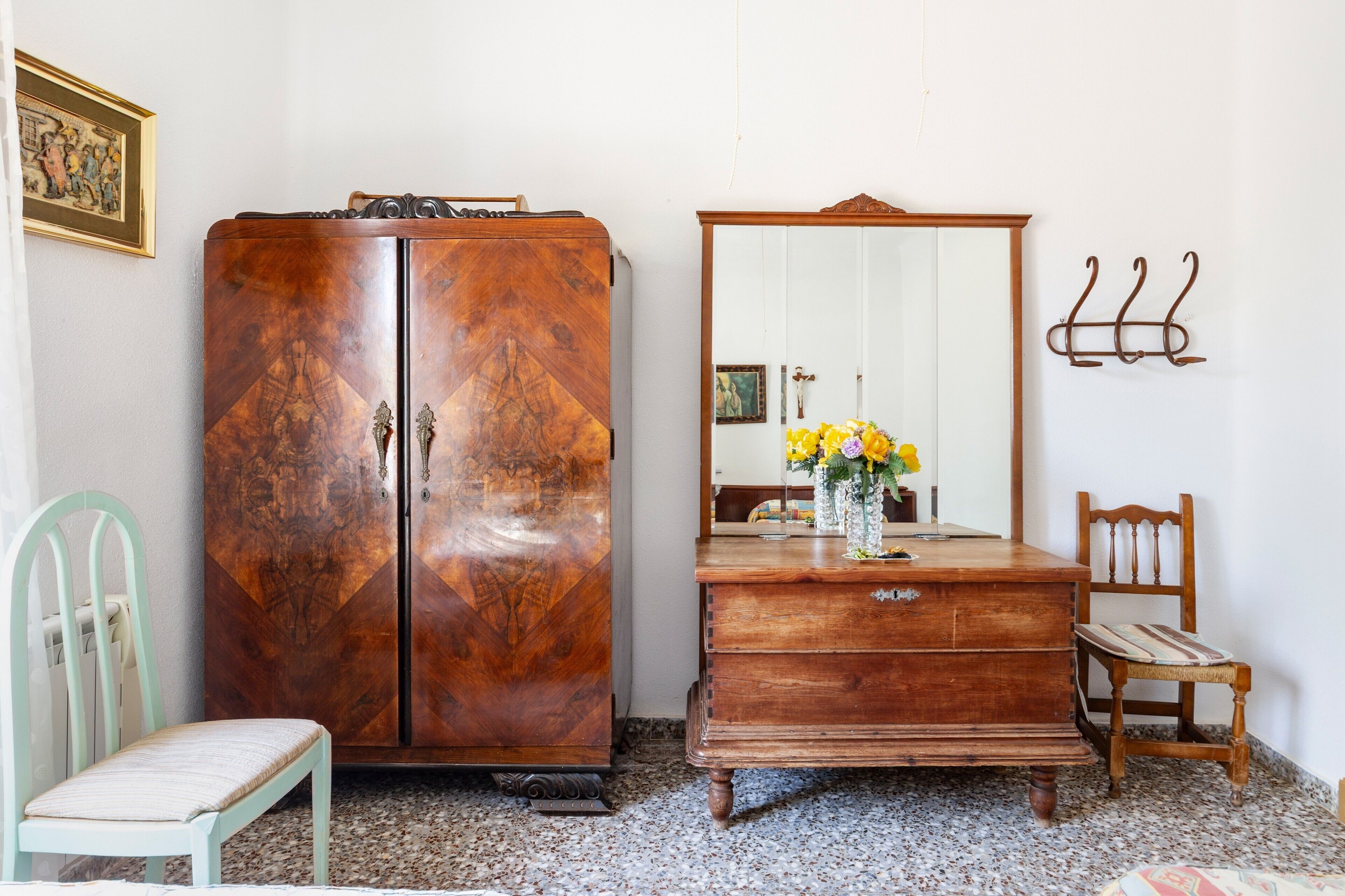 A room with a wooden wardrobe, a mirror, a chair, and a small table with flowers.