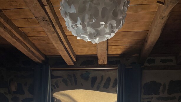 A rustic room with stone walls, wooden beams, a bed, a desk, and a view of an outdoor patio. - The House of Small Things (Lascelle)