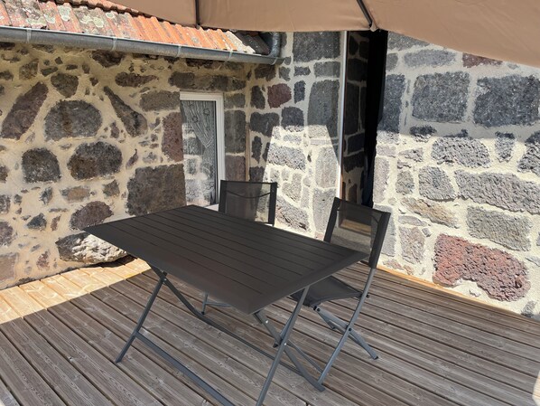 A stone exterior wall with a door and windows, a wooden deck, and a table with chairs. - The House of Small Things (Lascelle)