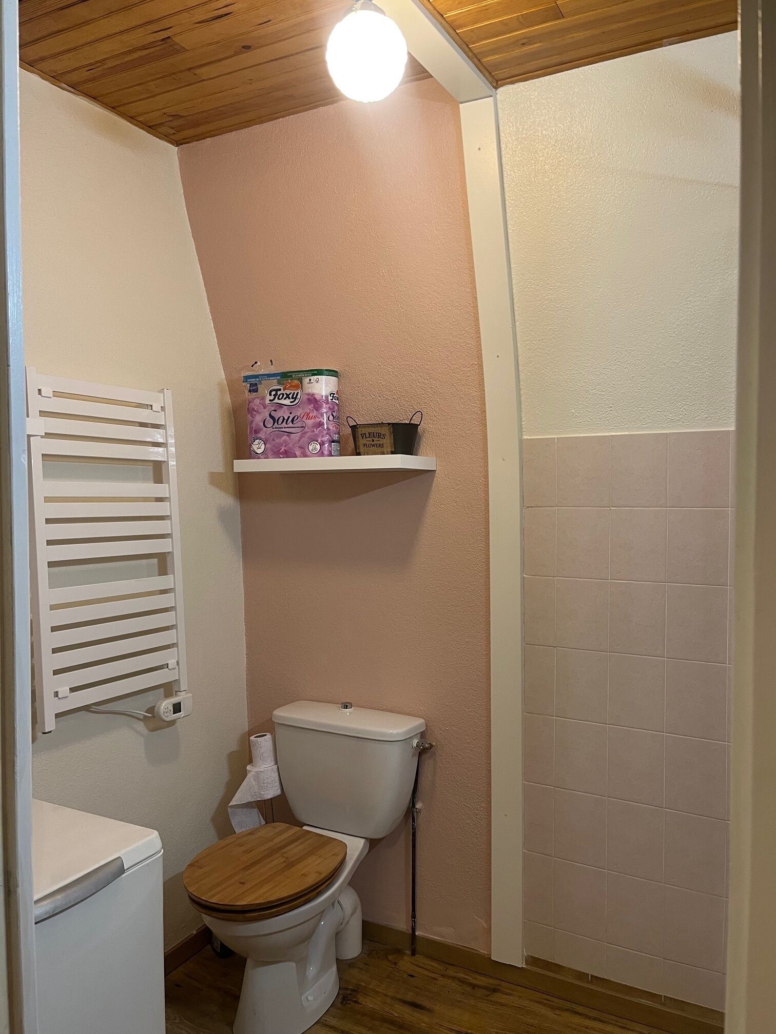 Une petite salle de bain avec des toilettes blanches, un plancher en bois, un radiateur blanc et une étagère contenant des articles de toilette.