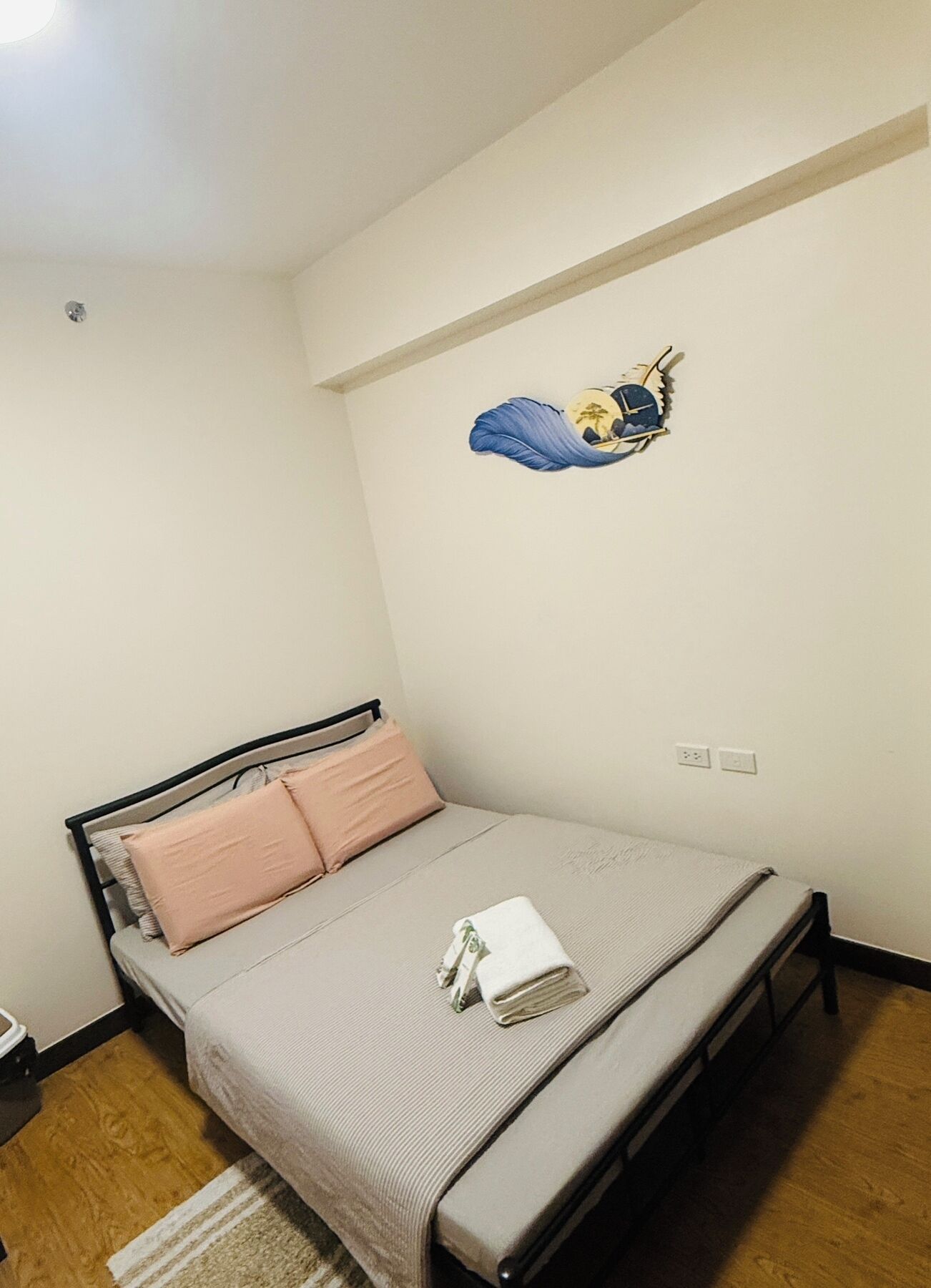 A small, neatly arranged bedroom with a single bed, a wall-mounted shelf with a decorative item, and a power outlet on the wall.