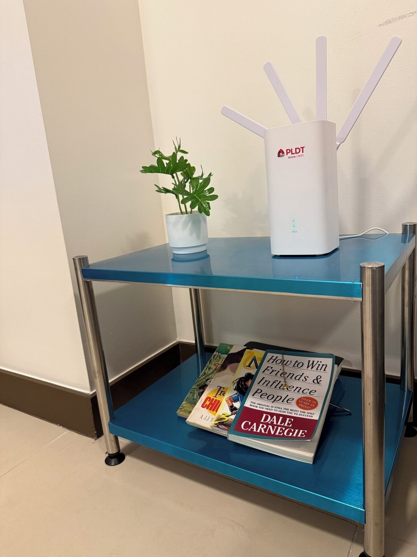 A small table with a white PLDT Home WiFi router, a potted plant, and a book titled "How to Win Friends & Influence People" by Dale Carnegie.