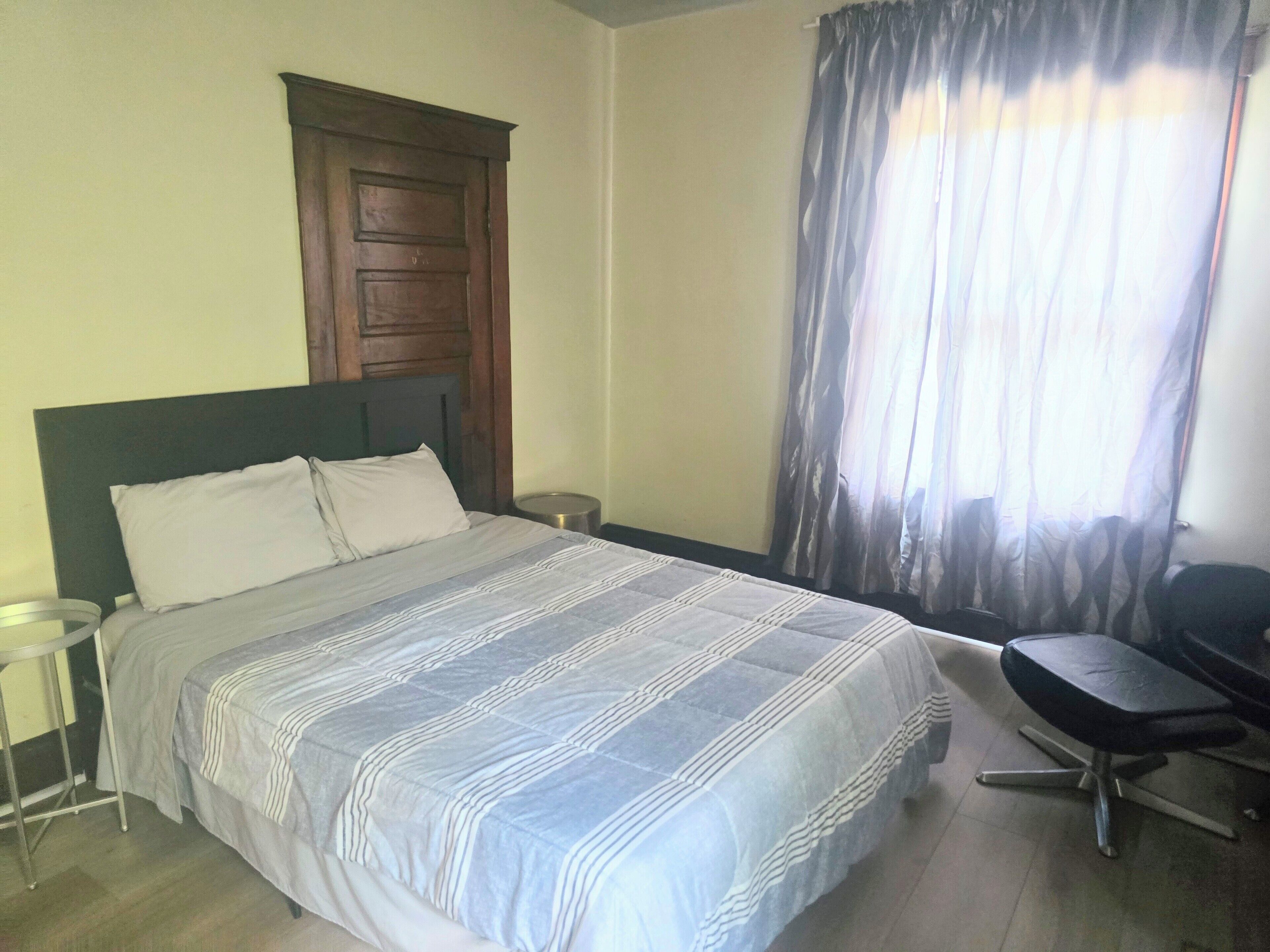 Ein Schlafzimmer mit einem Bett, einem hölzernen Kopfteil, einem Schreibtisch mit Stuhl, einem kleinen runden Tisch und einem Fenster mit Vorhängen.