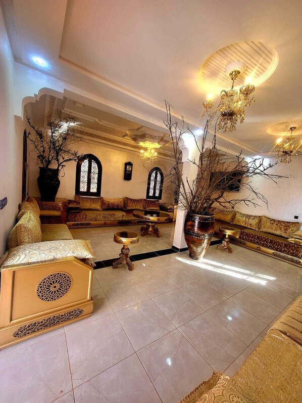 A spacious lounge area with a high ceiling, ornate chandeliers, and a central seating arrangement.