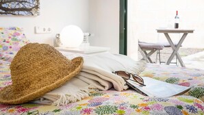 A bed with a straw hat, glasses, and a colorful floral blanket. There is a small table with a bottle and glasses, and a chair outside. - Villa Baix des Pujol: Modern Luxury & Relaxation in Mallorca (Petra)