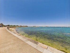 A coastal walkway with clear water, a stone barrier, and buildings in the background.