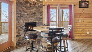 A rustic cabin interior with a stone wall, wooden ceiling, a round wooden table with chairs, a fireplace, and a large window with curtains.