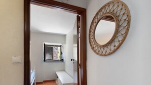 Un pasillo con marco de puerta de madera, un banco blanco, un ventanal con contraventanas y un espejo circular fijado a la pared.