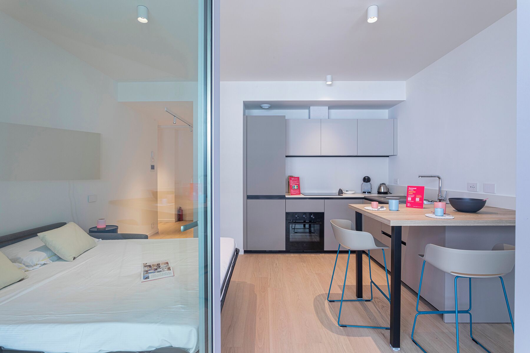 A modern, compact hotel room with a glass partition separating the bedroom from the kitchenette.