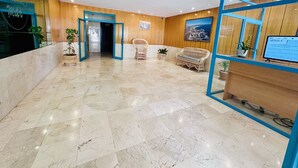 A hotel lobby with a marble floor, wooden walls, a reception desk, wicker furniture, and potted plants.
