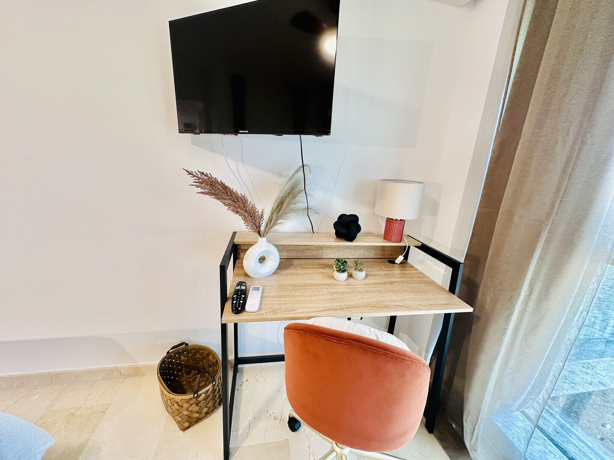 A small, modern workspace with a wooden desk, a mounted television, a lamp, and a chair.
