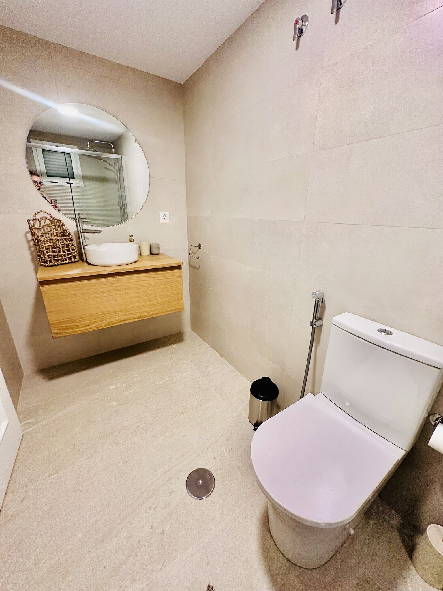 A modern bathroom with a white toilet, a wooden vanity, a round mirror, and a small trash bin.