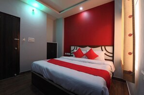 Habitación de hotel con cama, pared de acento roja, puerta de madera y un mueble pequeño.