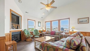 A spacious living room with a fireplace, large windows, and a ceiling fan. - Casa de Playa 4 Bedroom Home in Kill Devil Hills (Kill Devil Hills)