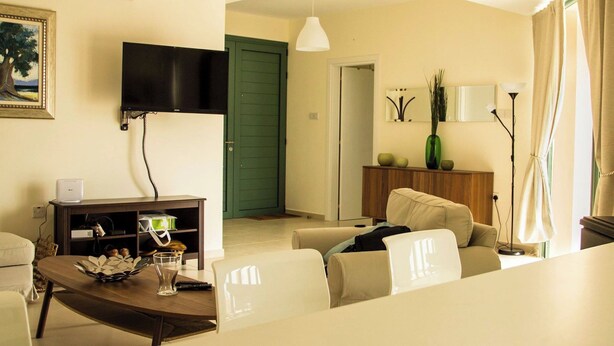 A modern living room with a flat-screen TV mounted on the wall, a dining table with chairs, a sofa, a cabinet with decorative items, and a green door in the background. - BELLEVUE VILLA. Completely private 3-bedroom villa with pool, gardens & orchard. (Anarita)