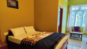 A neatly made bed with a floral bedspread and a quilted blanket, a wooden nightstand, and a chair near a window with curtains. - Gyalpo Homestay (Tawang)
