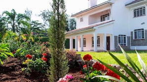 A white building with a covered porch and a well-maintained garden with various plants and flowers. - Spacious & Beautiful 5-bedroom Villa in Nairobi with WiFi (Nairobi)