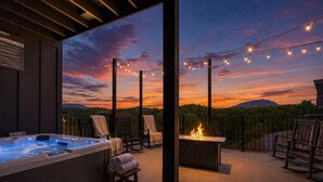 An outdoor patio with a hot tub, lounge chairs, a fire feature, and string lights against a sunset backdrop.