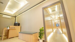 A reception area with a wooden desk labeled "RECEPTION," a potted plant, and a hallway leading to a bright interior. - Thrive Hotel (Bangkok)