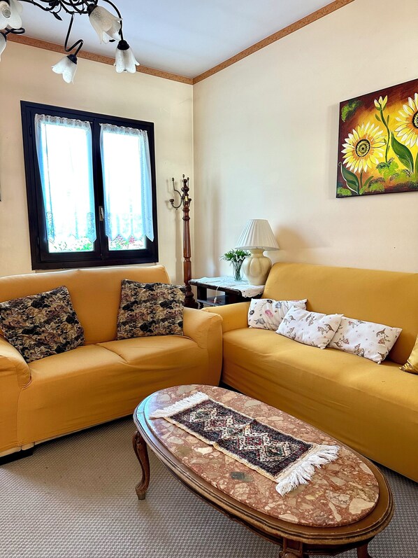 Ein Wohnzimmer mit einem gelben Sofa, einem gemusterten Teppich, einem hölzernen Couchtisch, einer Lampe und einem Sonnenblumenbild an der Wand.