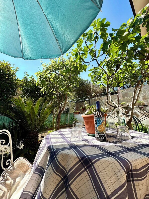 An outdoor seating area with a table covered by a plaid tablecloth, surrounded by greenery and a blue umbrella.
