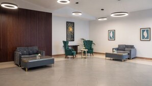 Un hall d’entrée spacieux avec un canapé gris, un fauteuil vert et un canapé bleu. On y trouve plusieurs luminaires au plafond et une grande œuvre d’art murale.