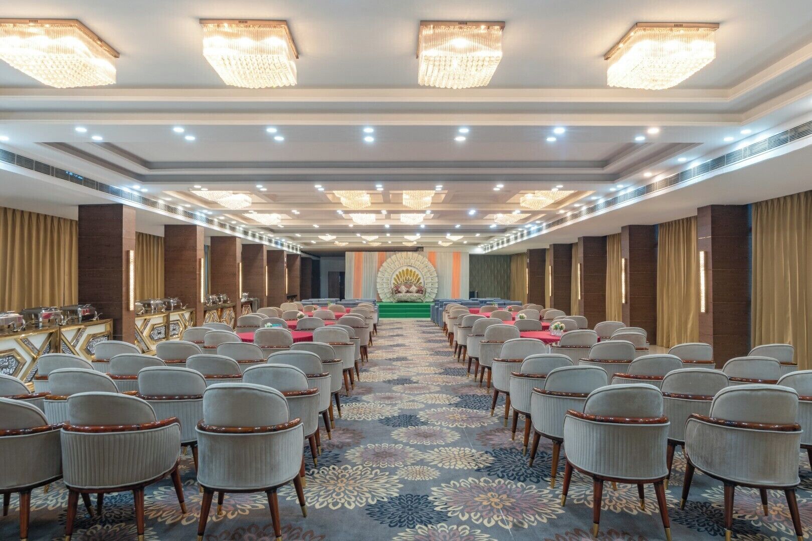 A conference room with rows of chairs, a stage with a green carpet, and a chandelier.