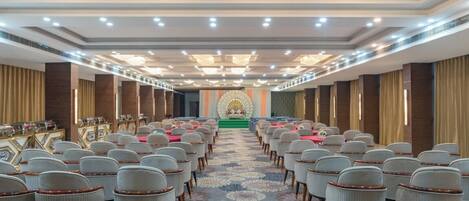 A conference room with rows of chairs, a stage with a green carpet, and a chandelier.