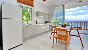 A modern kitchen with a refrigerator, stove, and sink. There is a dining area with a table and chairs. The room has large windows with a view of the ocean. - New Stunning Studio Watching Haad Yao Bay Sunset (Surat Thani)