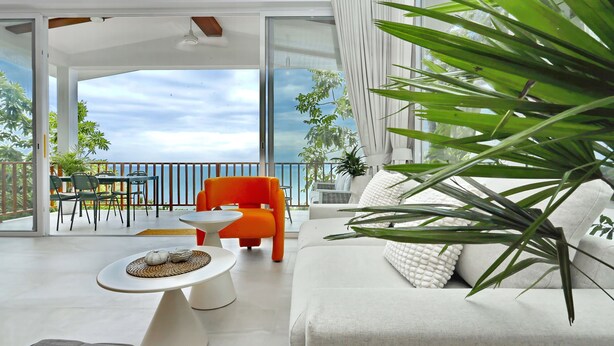 A modern living room with a white sofa, orange chair, and a small white table with decorative items. A balcony with a dining area and a view of the sea is visible. - New Stunning Studio Watching Haad Yao Bay Sunset (Surat Thani)