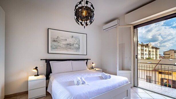 Una camera da letto moderna con un letto grande, un comodino, una lampada da notte, un quadro incorniciato al muro e vista sulla città da un balcone.