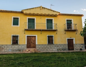 A yellow building with a tiled roof, two balconies with metal railings, and a brick base.