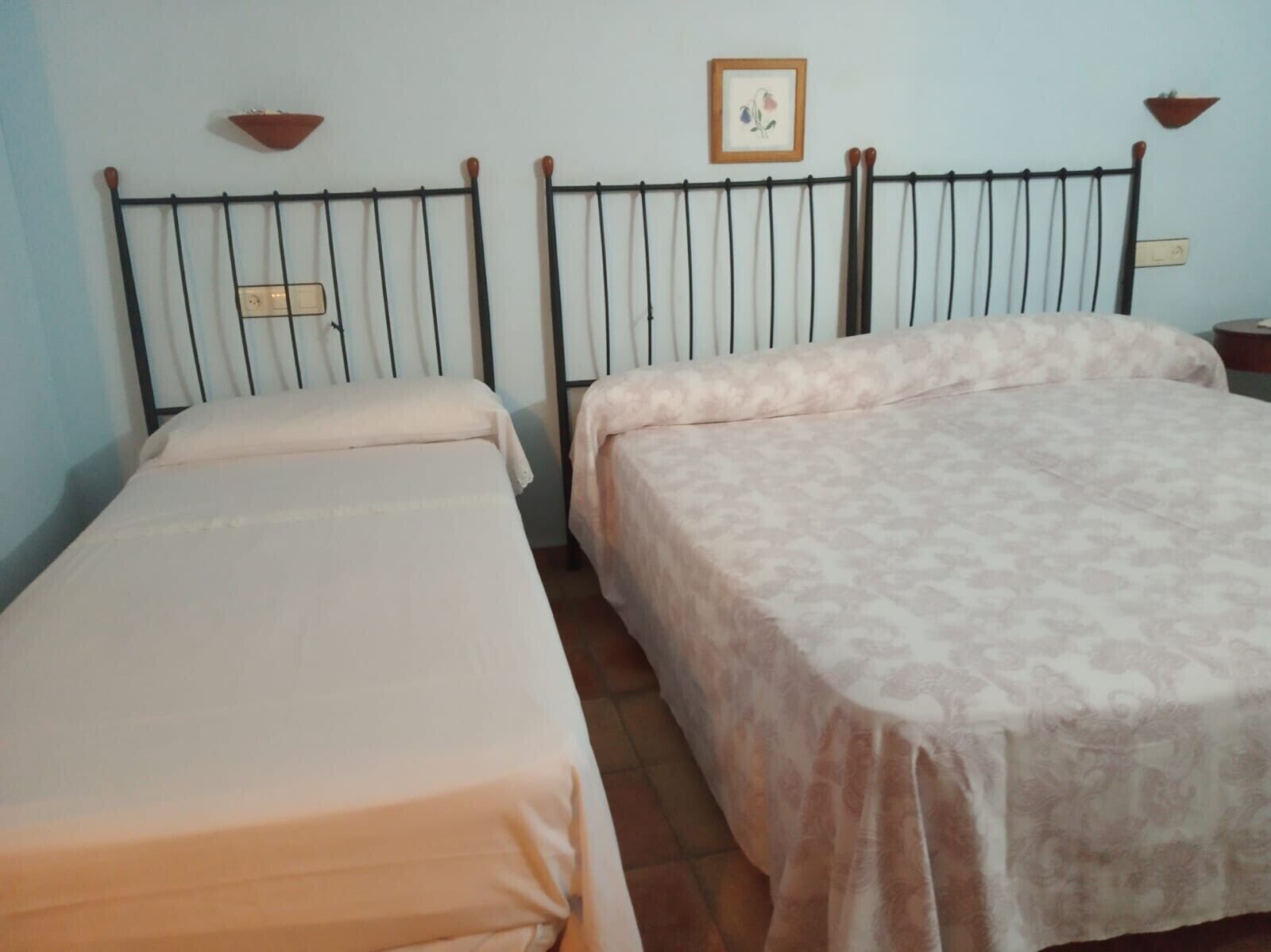 A room with two beds, a wall-mounted shelf, a framed picture, and a bedside table.