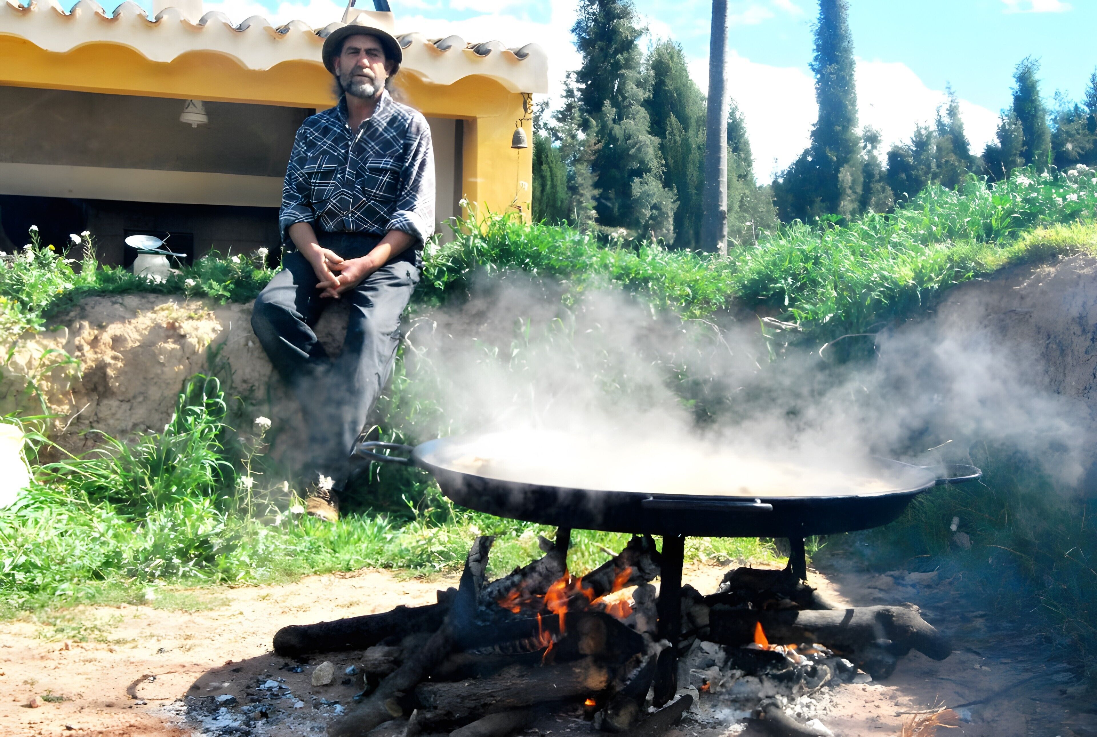 A person sitting next to a large outdoor cooking stove with a flat cooking surface, surrounded by greenery and a building in the background.
